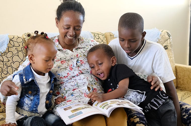 sos-mamma i blommig skjorta och ler sitter i soffa med tre nyfikna barn och en bok i knät ena barnet pekar i boken