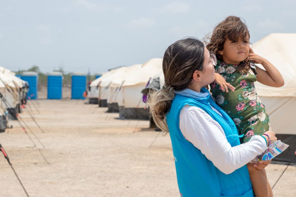En kvinna lfyter upp ett barn framför en bakgrund som visar flyktingtält.