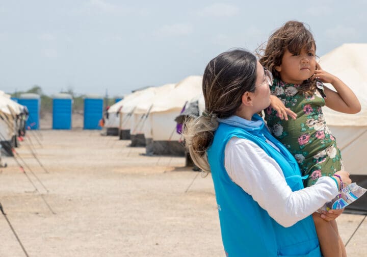 En kvinna lfyter upp ett barn framför en bakgrund som visar flyktingtält.
