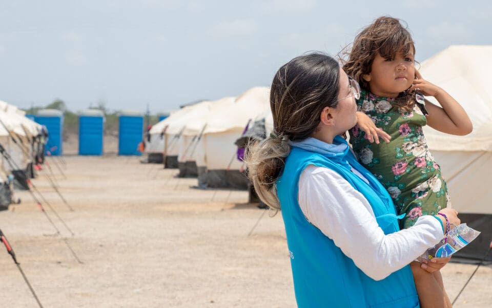 En kvinna lfyter upp ett barn framför en bakgrund som visar flyktingtält.